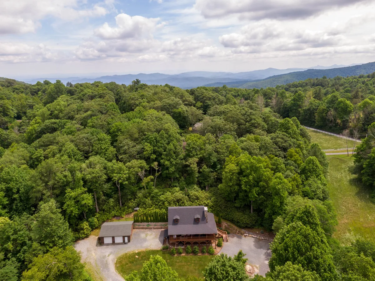 A wide view of the main house surrounded by lush trees, with the mountains providing a dramatic backdrop and the driveway leading up to the entrance. The setting is tranquil and immersed in nature, inviting guests to explore the outdoors and nearby attractions.