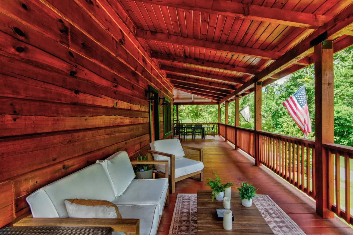 A cozy wooden deck furnished with comfortable white sofas and potted plants, framed by the American flag and surrounded by greenery. The space evokes a welcoming atmosphere for socializing, watching sunsets, or starting the day with coffee outdoors.