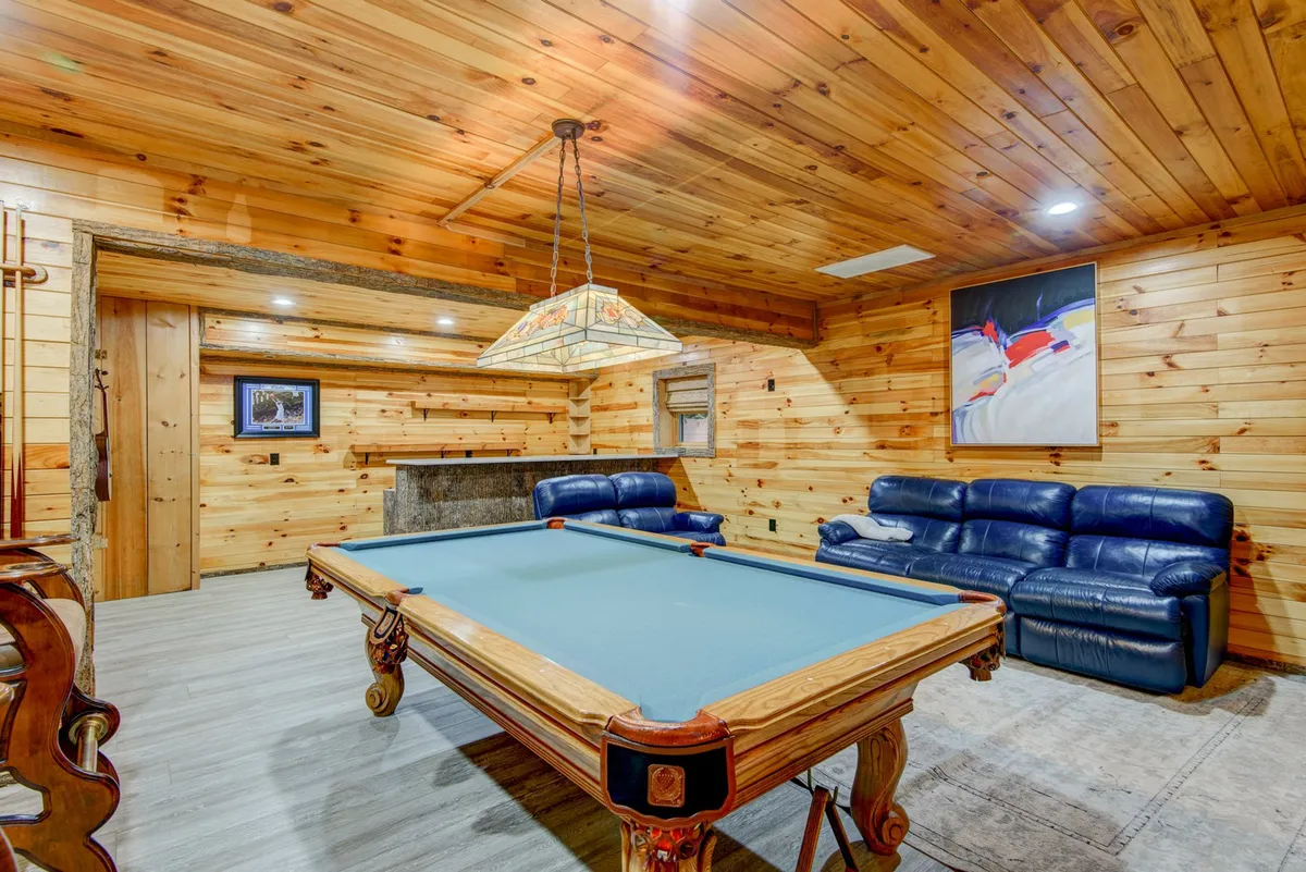 A cozy, dedicated game room featuring a pool table, leather couch, warm wood walls, and a stylish hanging light. The room is set up for family tournaments and social gatherings after a day of outdoor fun.