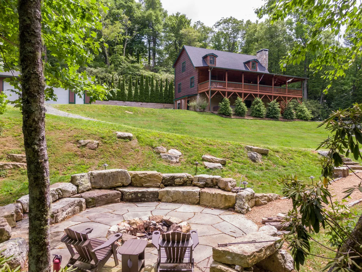 A wide view showing the house, the fire pit area in use, and people relaxing on the porch. Surrounded by trees, the image captures both the house and its vibrant outdoor living spaces, emphasizing the seamless transition from inside to outside living.
