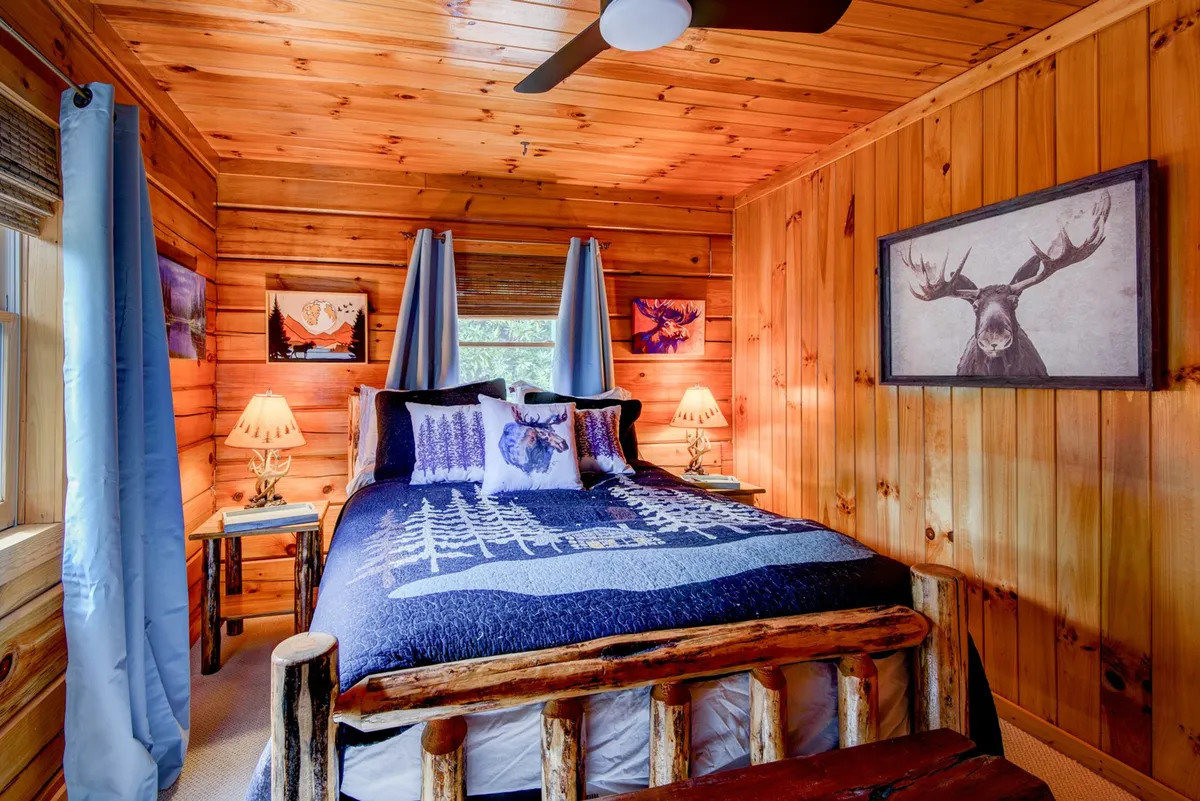 A warm bedroom with a wooden bed frame, blue bedding adorned with tree patterns, framed animal artwork, and bedside lamps. The window allows natural light to fill the space, enhancing the inviting and peaceful atmosphere.