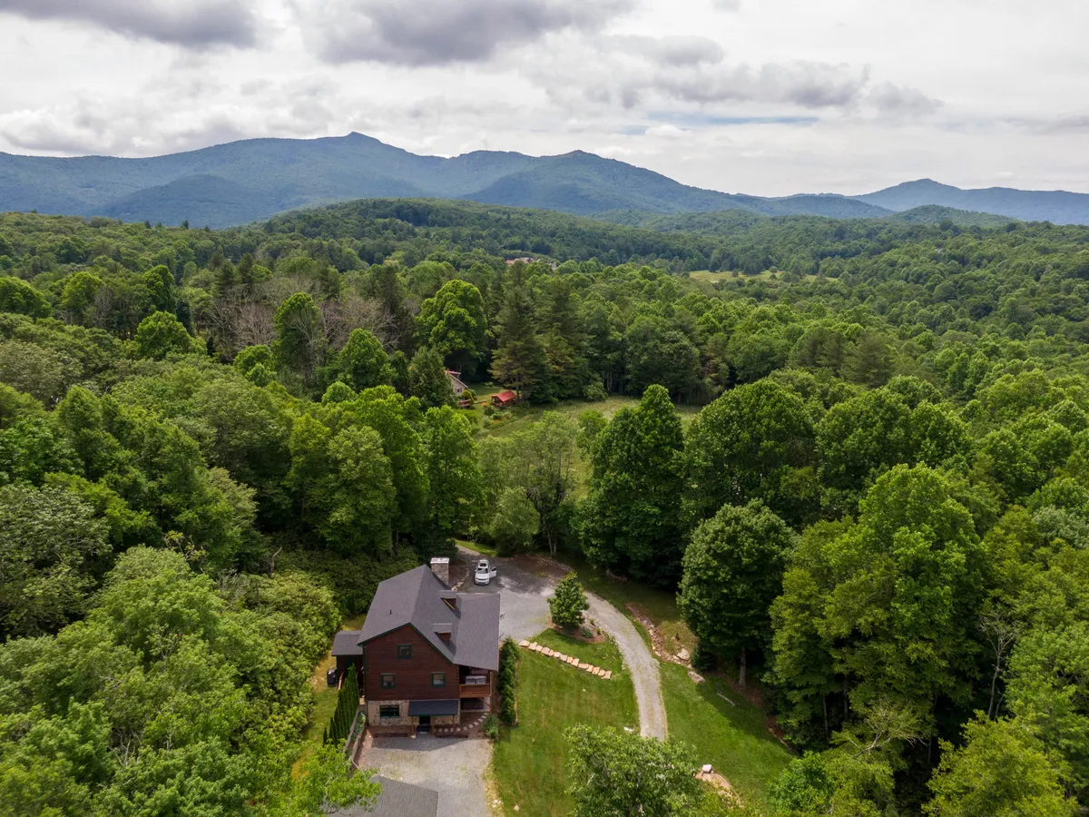 This image features the house nestled among lush trees with a scenic mountain backdrop, immediately evoking the spirit of outdoor adventure in the Blue Ridge Mountains. The composition showcases proximity to nature and hints at nearby hiking and exploration opportunities.