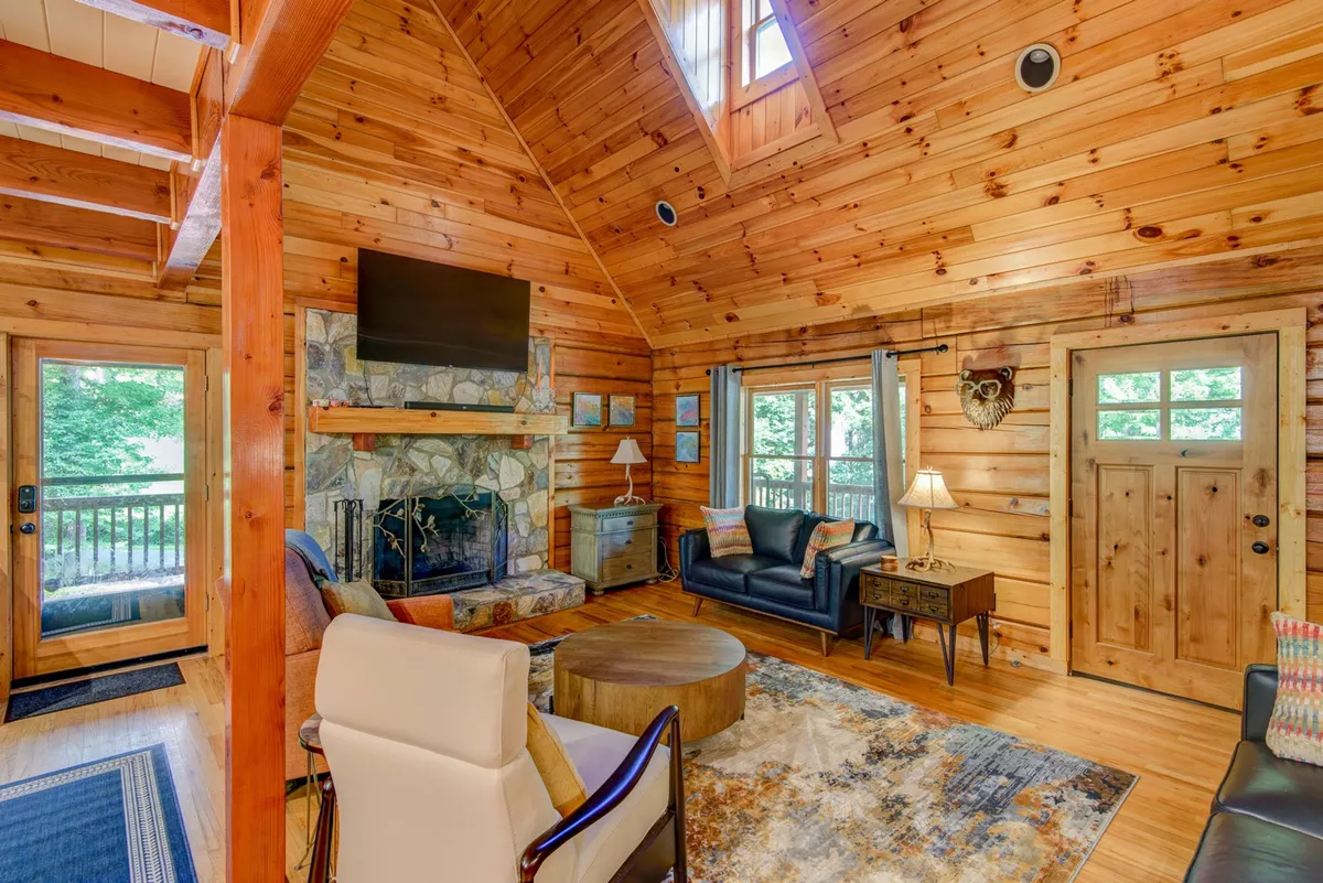This image showcases a spacious open-concept living area with a prominent stone fireplace, large windows letting in natural light, wooden beams across the ceiling, comfortable seating, and artwork on the walls. The arrangement is ideal for family gatherings, reading, or cozying up by the fire.