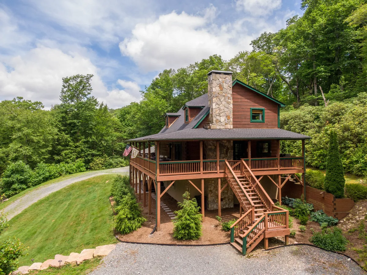 This image showcases the charming exterior of Rhododendron Rendezvous, featuring a stone chimney, wraparound porch, and lush greenery all around. The inviting porch offers views of the surrounding mountain landscape, perfect for morning coffee or sunset relaxation.