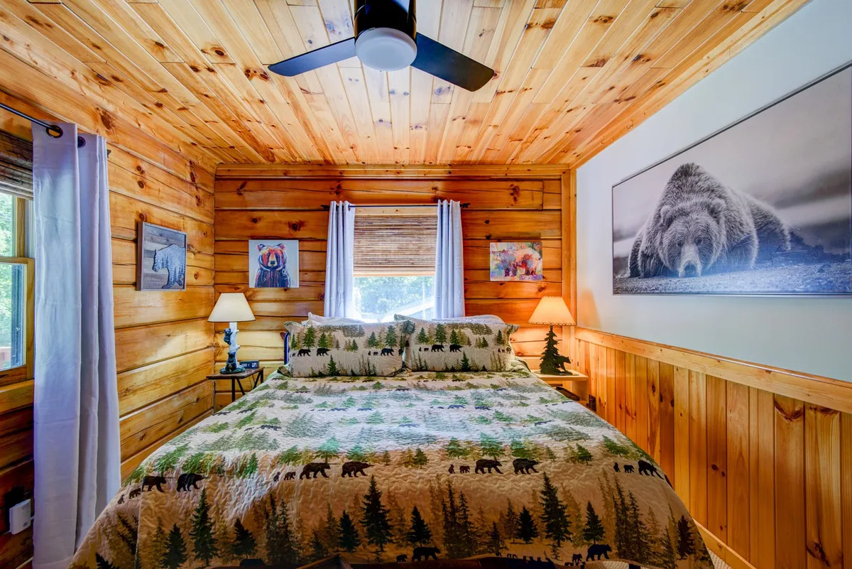 A cozy bedroom featuring a bed with forest-themed bedding, warm wooden wall paneling, a framed bear photograph above the bed, a nightstand with a lamp, and a window with curtains letting in natural light. The room exudes a sense of calm, inviting guests to relax and unwind.
