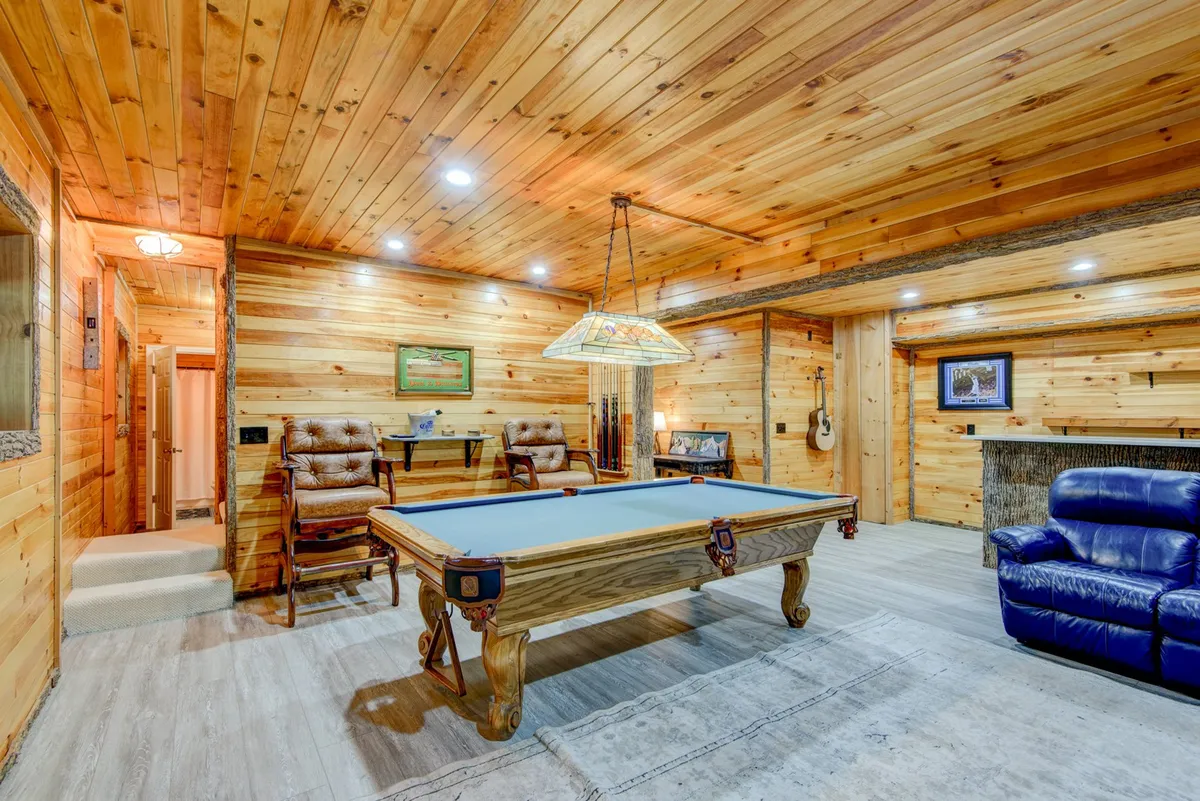 A dedicated billiards room with a pool table as the centerpiece, complemented by a reclining chair, wood-paneled walls, and a hanging light fixture overhead. The casual, welcoming vibe is perfect for games and socializing.