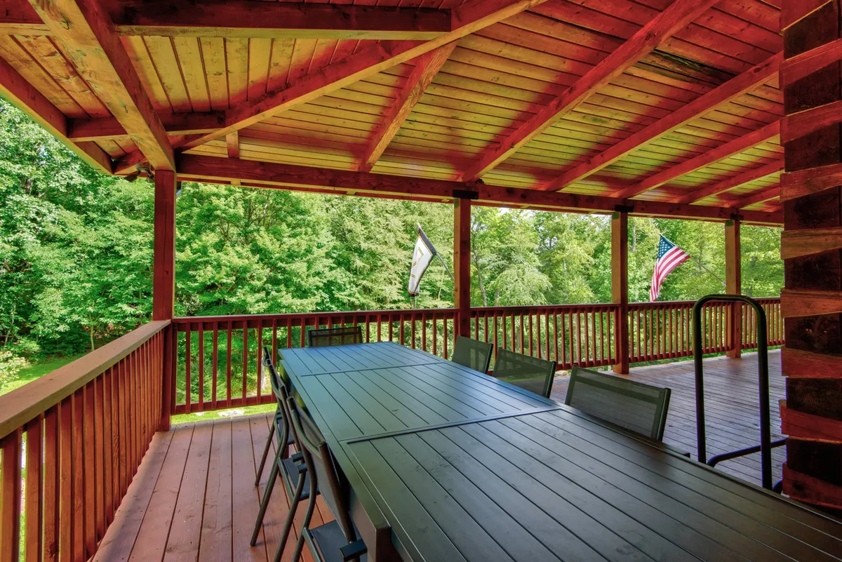 A spacious outdoor dining area featuring a large wooden table set beneath sturdy wooden beams, surrounded by lush greenery and adorned with festive flags. The setting is perfect for hosting group meals, celebrations, and relaxed conversations, all with a backdrop of natural beauty.