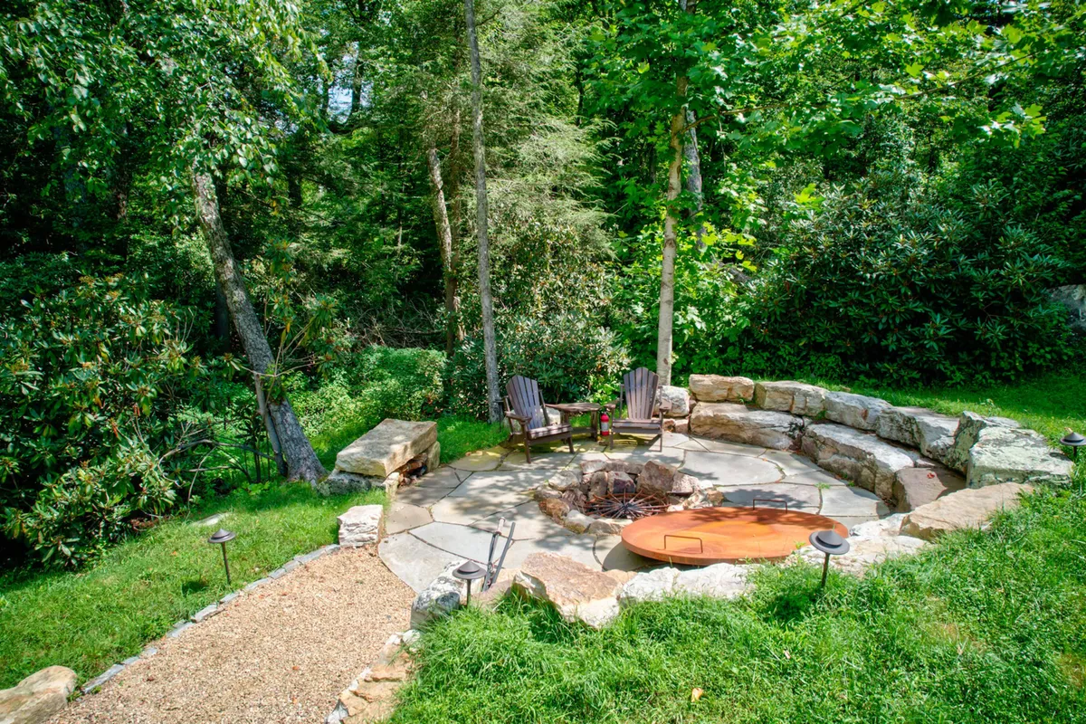 A stone fire pit with circular stone seating, nestled against a wooded backdrop. The area is designed for small gatherings, storytelling, and enjoying the warmth of a crackling fire under the stars, with nature enveloping the space.