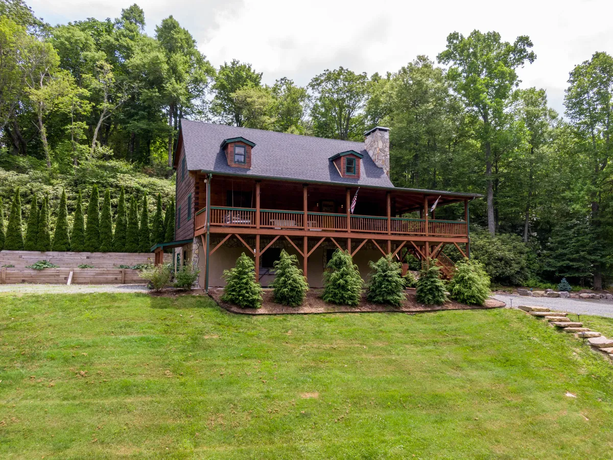 The image showcases the welcoming wooden porch of Rhododendron Rendezvous, complemented by a stone chimney and manicured lawn, all surrounded by lush trees. The porch features comfortable seating, inviting guests to relax and enjoy the outdoors. The natural setting extends the living space and highlights the seamless transition between the home and its scenic mountain surroundings.