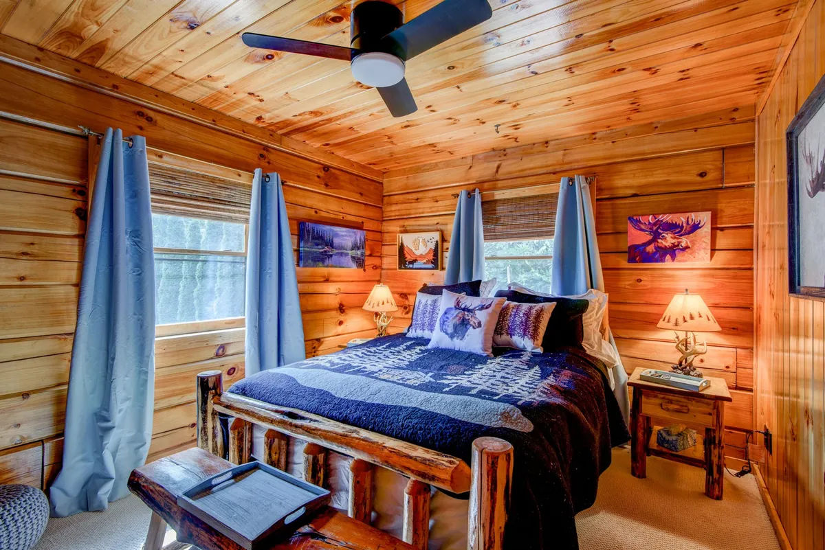 A peaceful bedroom with a bed adorned with decorative pillows, a wooden ceiling, blue curtains, bedside lamps, and artwork on the walls. The ambiance is tranquil and warm, offering a perfect setting for restful sleep and relaxation.