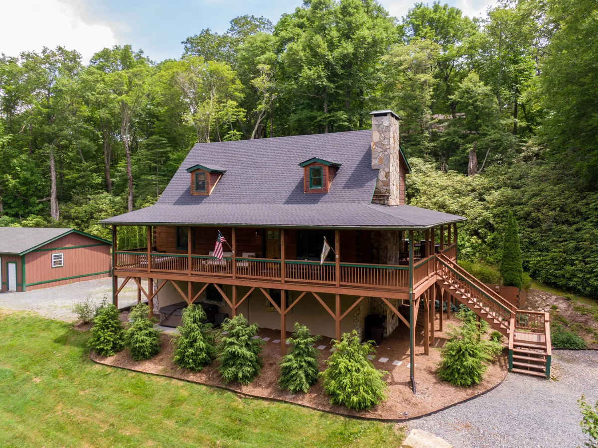 The image showcases a charming log cabin nestled amid lush greenery, with a prominent stone chimney, inviting front porch, and warm sunset lighting. The setting evokes tranquility and privacy, perfect for cozy outdoor gatherings and relaxation.
