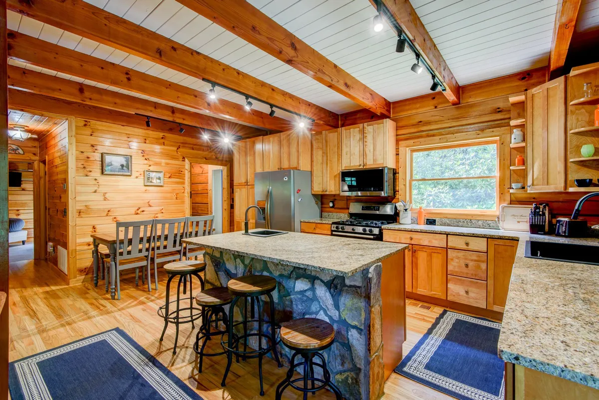 A warm, communal kitchen and dining space with a prominent granite island, visible wooden beams, stainless steel appliances, and a cozy dining area. The layout is open, designed for group cooking and casual family gatherings, with clear sightlines and comfortable seating.