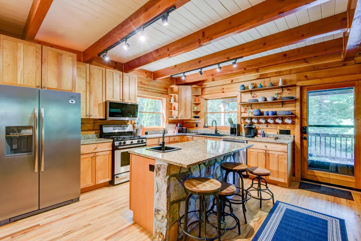 A beautifully designed kitchen featuring a central island with a stone base, stainless steel appliances, wooden cabinetry, and open shelving displaying dishware. Large windows flood the space with natural light, enhancing the welcoming ambiance ideal for cooking and socializing.